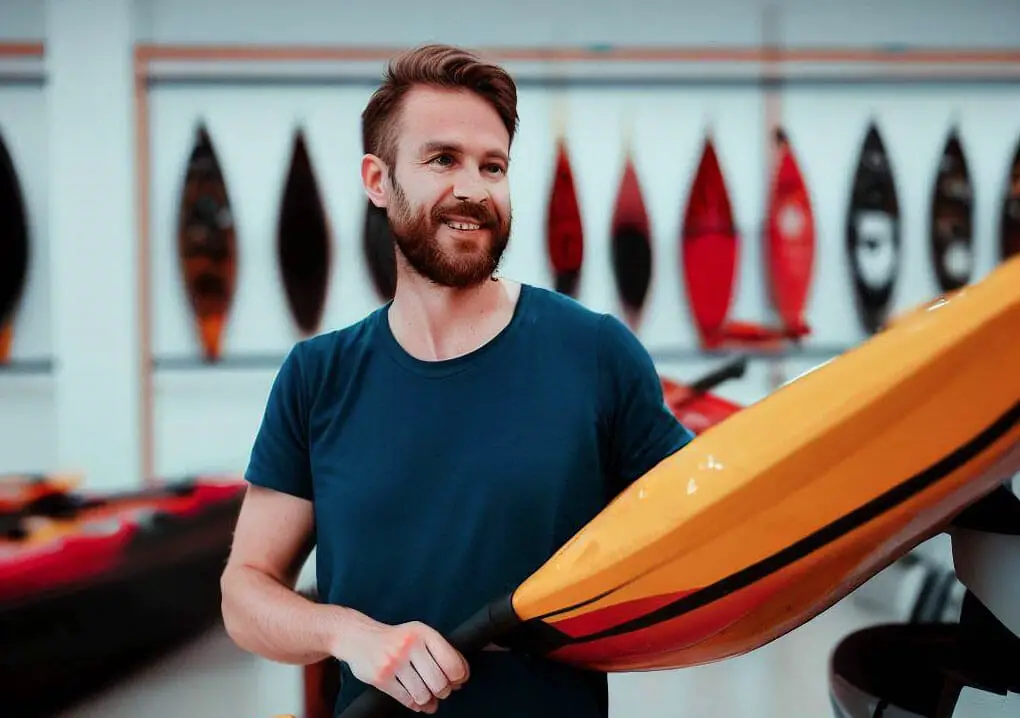 A man with a beard standing next to a kayak, referencing the "Kayak Buying Guide".